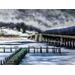 An icy, frozen river with a deck extends onto it. A bridge on right crosses the other side, a riverbank of Winter grasses with a snowy hill.